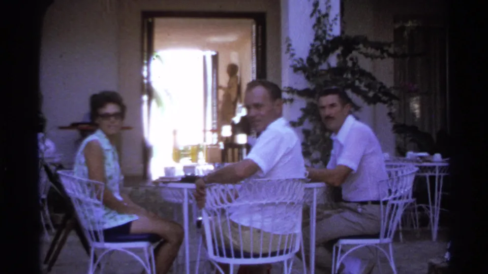 1974 Cabo San Lucas patio scene with three adults seated at a white metal table, looking toward the camera.