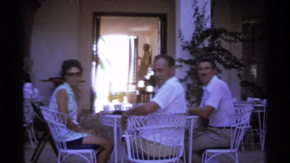1974 Cabo San Lucas home-movie frame of three adults seated outdoors at a patio table with drinks nearby.