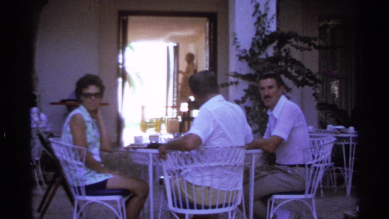 In Cabo San Lucas home-movie footage from 1974, three adults sit at a white wicker patio table under shaded cover.
