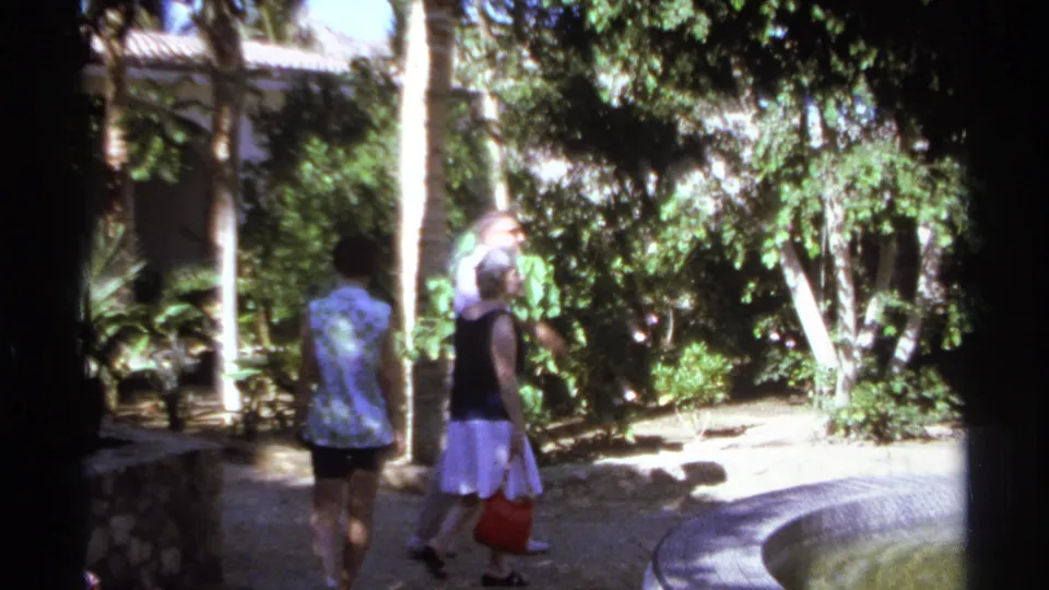 In the 1974 Cabo San Lucas garden scene, two adults walk past palms and dense greenery beside a stone fountain.