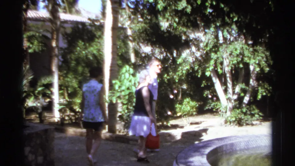 In 1974 Cabo San Lucas footage, adults walk through a shaded tropical garden, with one woman carrying a red handbag near a stone path.