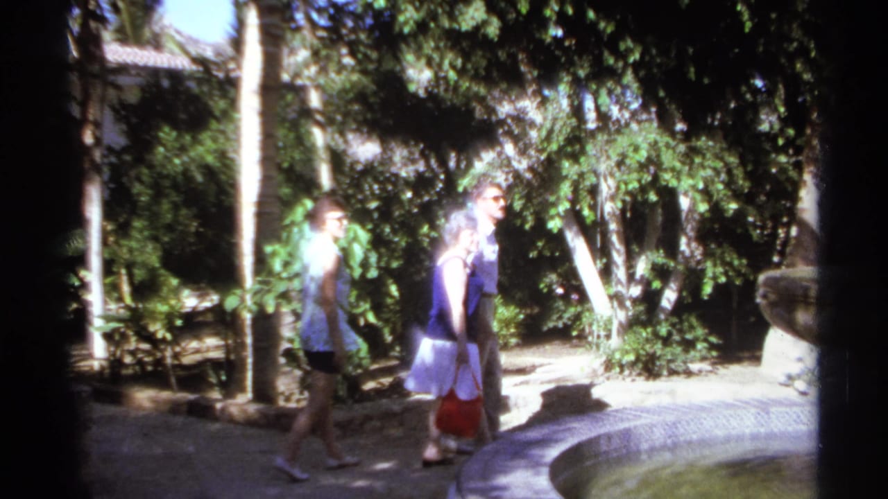 Three adults continue along a garden path near trees and a curved fountain edge.