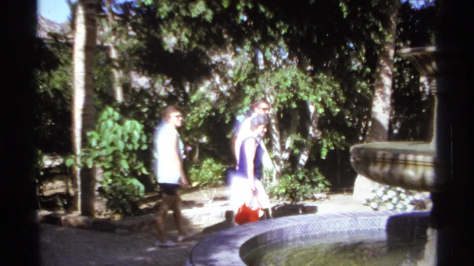 Adults in casual clothing walk beside a curved fountain basin in a sun-dappled tropical garden, consistent with the 1974 Cabo San Lucas metadata.