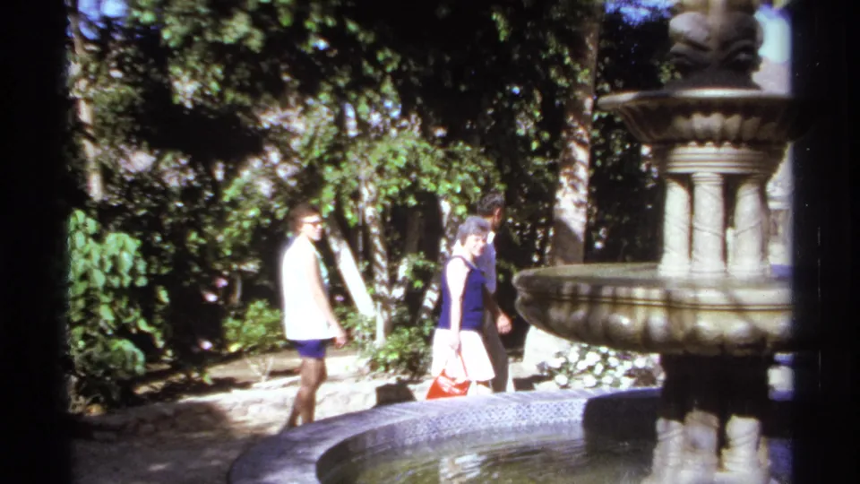 Adults walk past a tiered decorative fountain in a Cabo San Lucas garden, with palms and shaded foliage behind them.