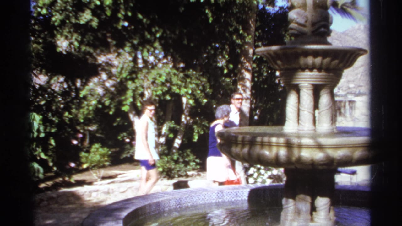 Adults walk through a garden behind a large stone fountain.