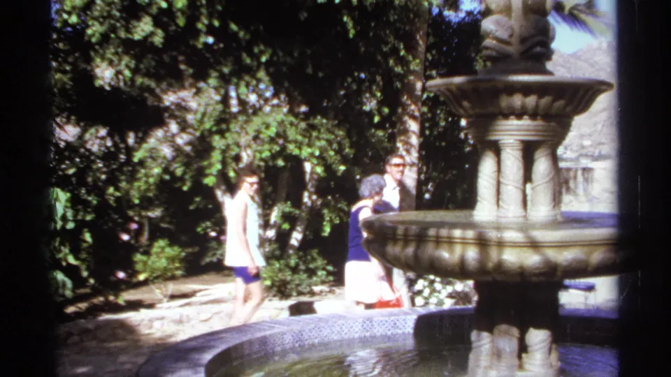 In 1974 Cabo San Lucas home-movie footage, adults pass behind an ornate stone fountain in a sunny tropical garden.