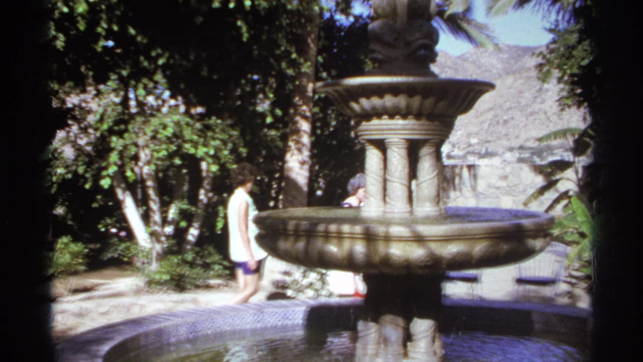 A man in a light shirt walks beside a large fountain in a garden.