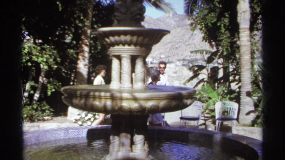 Adults gather and pass near an ornate stone fountain in bright 1974 Cabo San Lucas garden footage.