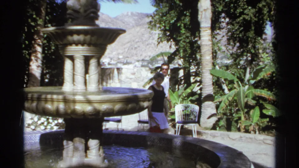 A man in sunglasses is visible beside the fountain in a sunlit Cabo San Lucas garden, with palms and chairs nearby.