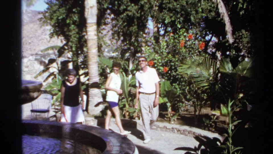 In supplied 1974 Cabo San Lucas home-movie footage, three adults walk on a sunlit garden path beside a circular fountain and dense tropical plants.