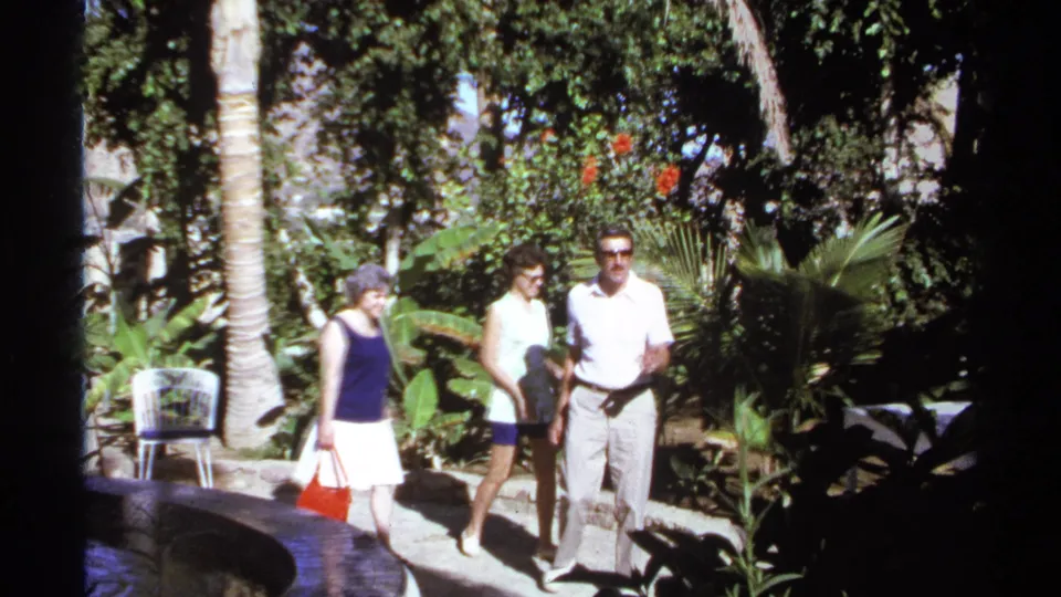 A woman carrying a red bag, a young woman in shorts, and a man in a white shirt pass a fountain edge in the supplied 1974 Cabo San Lucas garden scene.