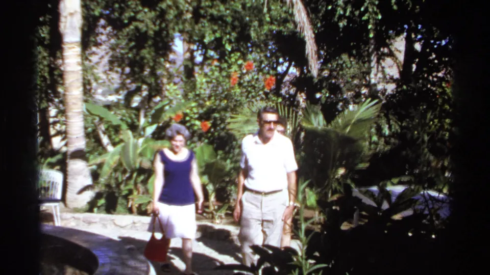 A woman with a red bag and a man in sunglasses walk closer to the camera in the supplied 1974 Cabo San Lucas tropical garden footage.