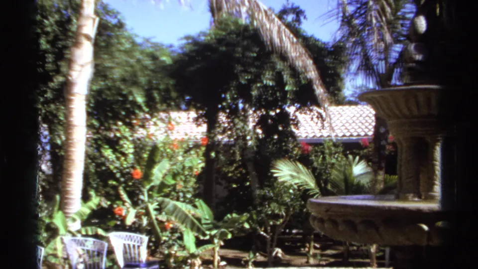The final frame centers the garden setting, with a stone fountain, white metal chairs, palms, and thick foliage in 1974 Cabo San Lucas footage.