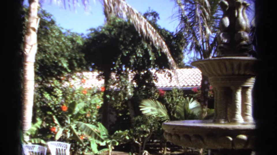1974 Cabo San Lucas garden view with a tiered stone fountain, tropical foliage, red flowers, tiled roof and blue sky.