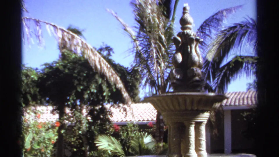 1974 Cabo San Lucas home-movie frame centered on a decorative garden fountain with palms, vines, roof tiles and sunlit foliage.