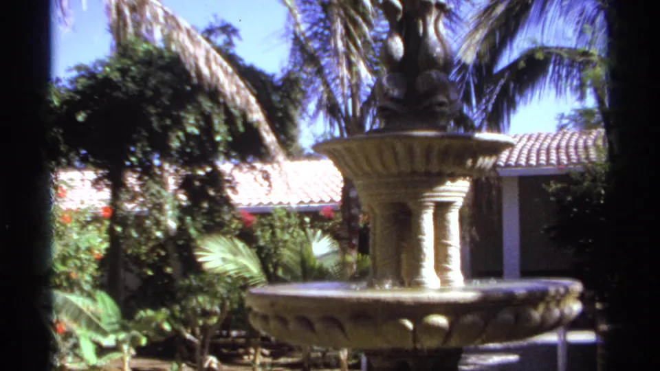 1974 Cabo San Lucas garden shot with the fountain at right, tropical greenery, red flowers, tiled roof and bright sky.