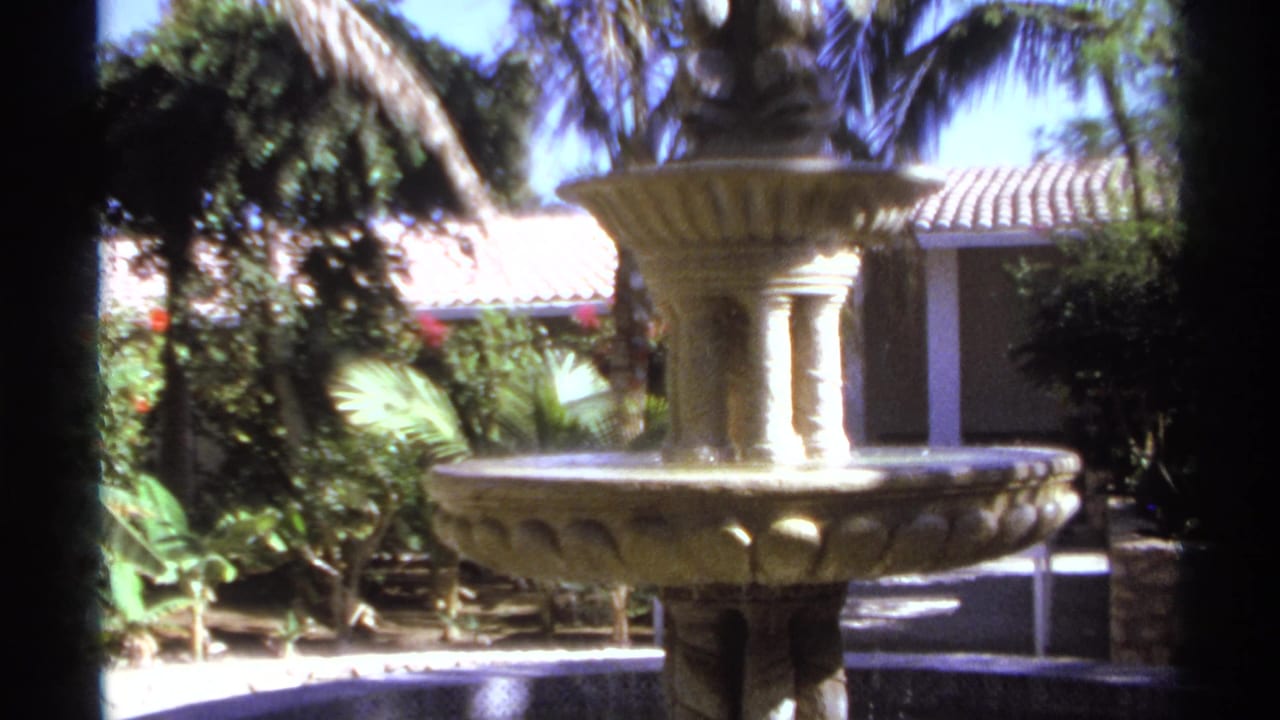 1974 Cabo San Lucas home-movie frame showing a two-tier stone fountain, palm foliage, shrubs, and a terracotta-roofed building.