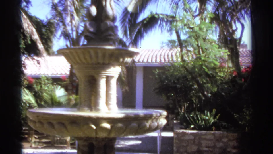 1974 Cabo San Lucas home-movie still showing the fountain lower basin, tropical greenery, stone planter, and tiled roofline.