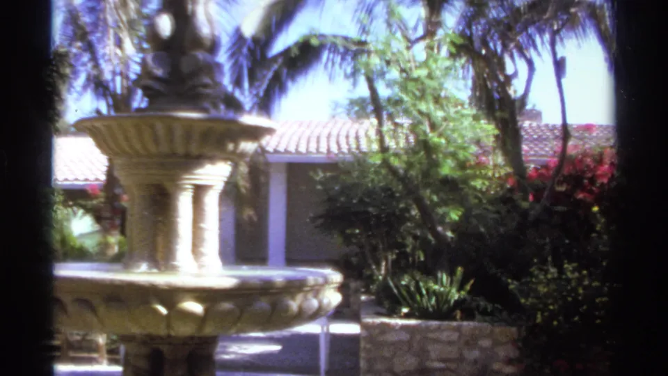 1974 Cabo San Lucas frame with the fountain partly left of center, backed by palms, red flowering shrubs, and a terracotta roof.
