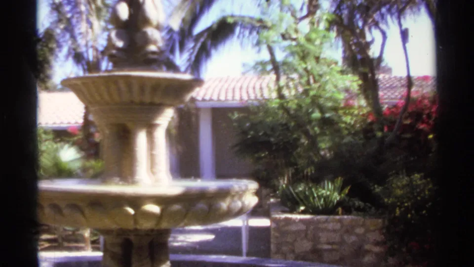 1974 Cabo San Lucas frame centered on a stone fountain set among palms, shrubs, low stone walls, and a tiled-roof courtyard building.