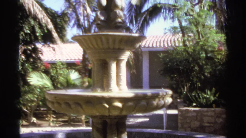 1974 Cabo San Lucas home-movie frame showing a tiered stone fountain with visible water, tropical foliage, and courtyard stonework.