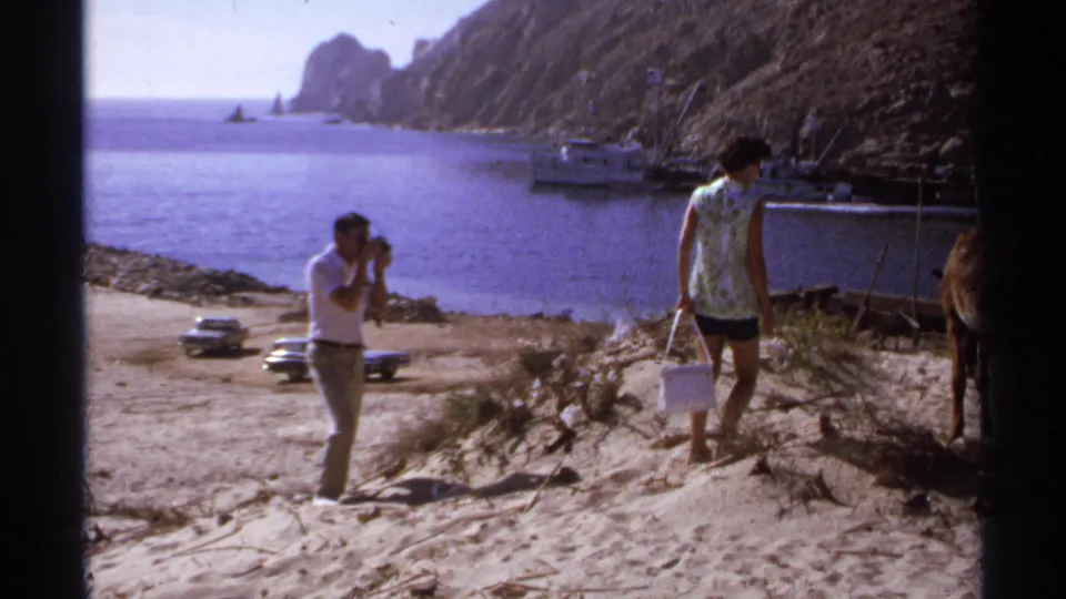 1974 Cabo San Lucas frame with a man using a camera as a woman carrying a bucket walks near a donkey by the harbor.