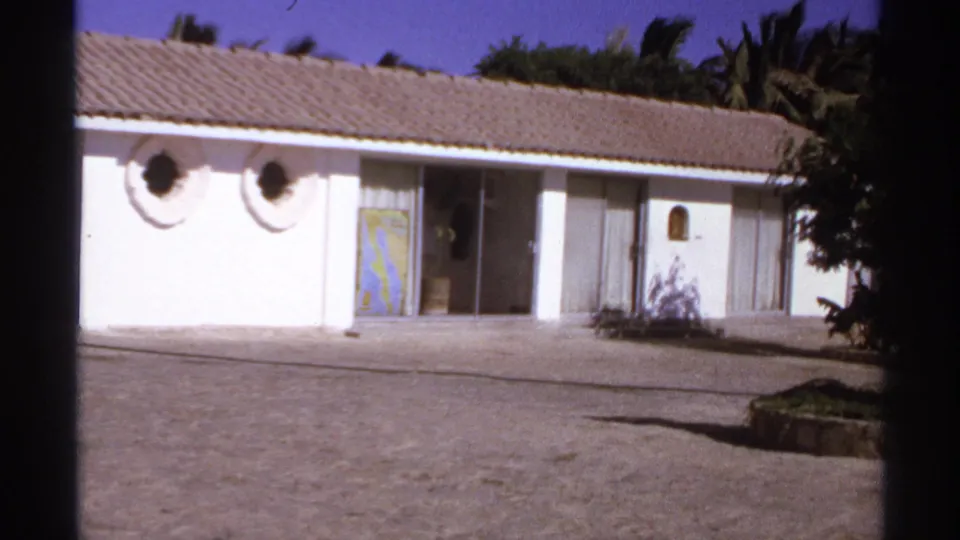 1974 Cabo San Lucas resort-area frame showing a white low building with circular windows, tiled roof, open doorway, pavement, and palms.