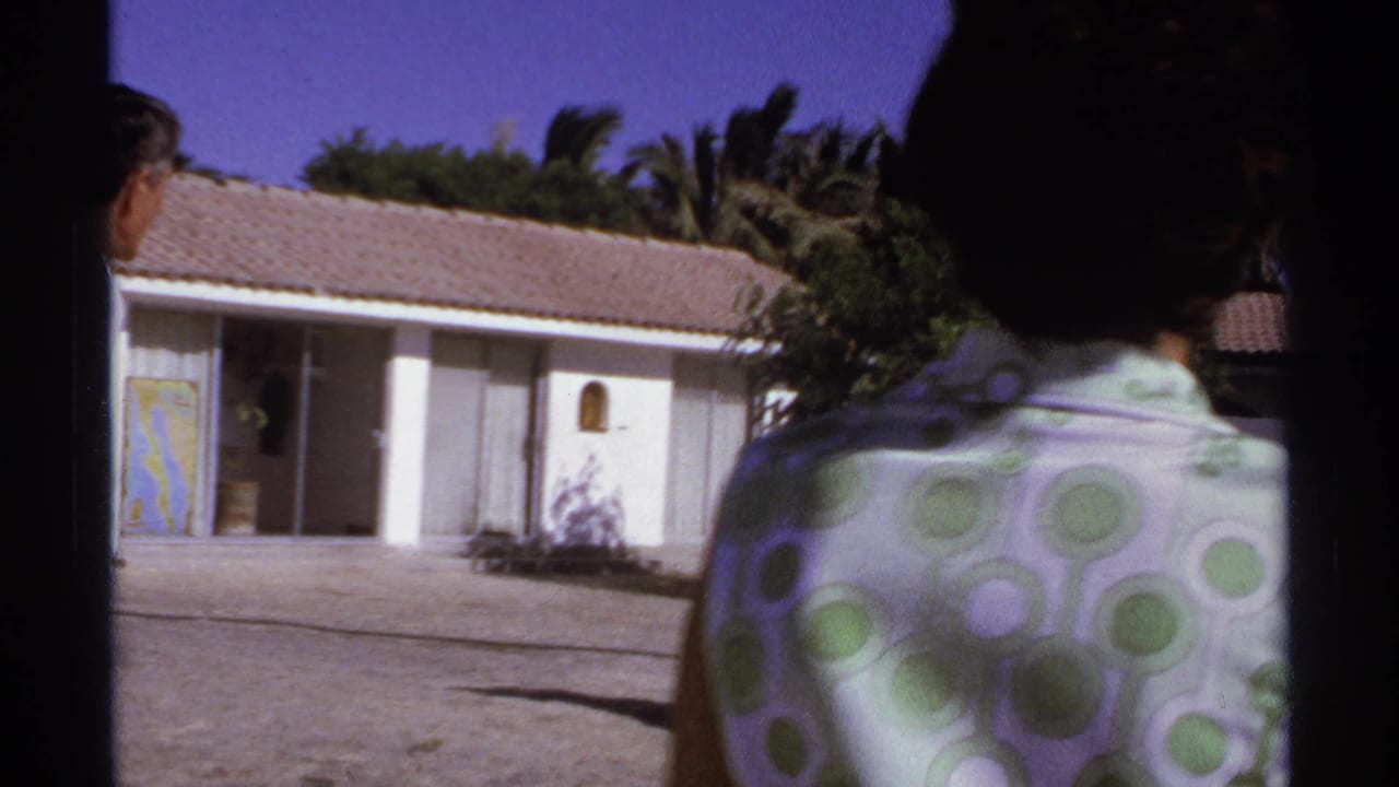 1974 Cabo San Lucas home-movie frame of a woman in a green polka-dot dress walking toward a white stucco house with palms behind it.