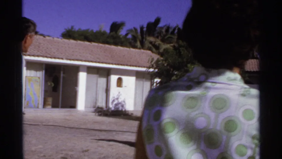 1974 Cabo San Lucas home-movie frame of a woman in a green polka-dot dress walking toward a white stucco house with palms behind it.