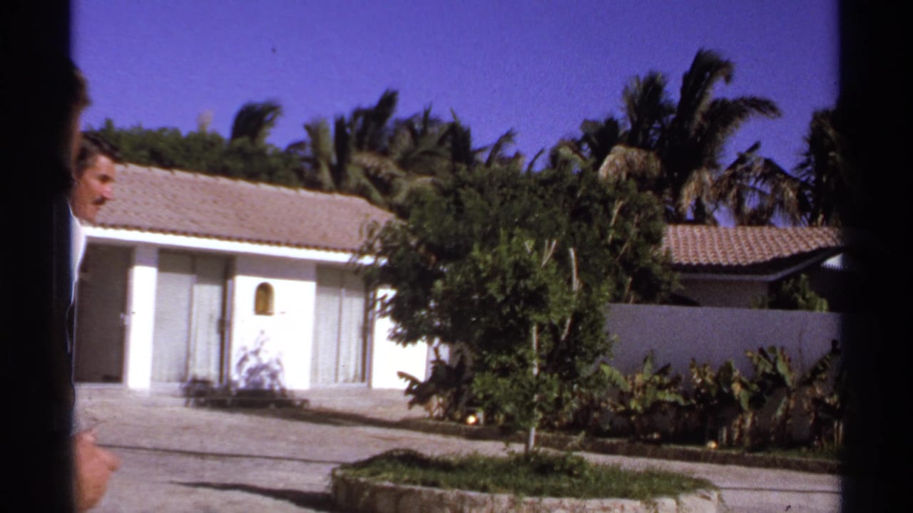 Man partially visible near a white stucco house, with palms and tropical plants in the background.