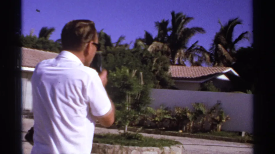 1974 Cabo San Lucas home-movie frame of a man in a white polo shirt and sunglasses walking beside a stucco wall and palm trees.