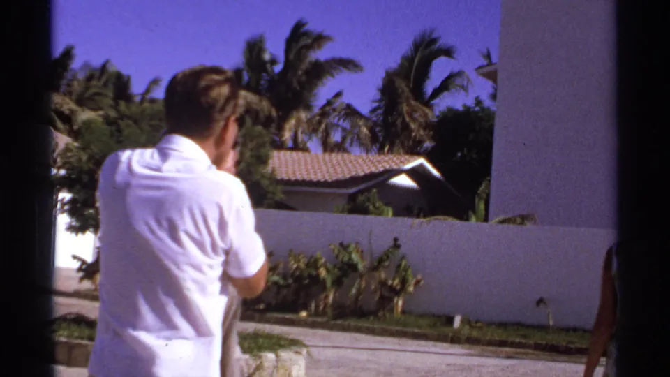 1974 Cabo San Lucas frame of a man looking across a sunny tropical residential setting with stucco wall, tile roof, and palms.