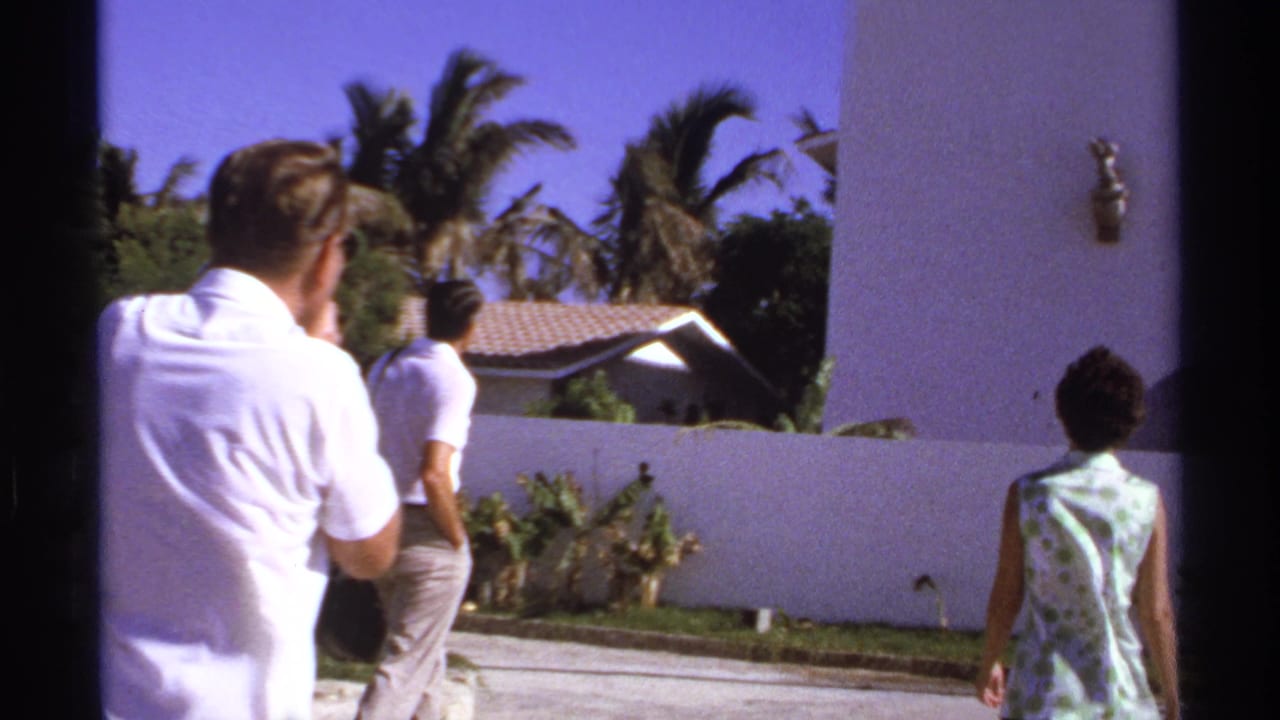 Adults walk beside a white stucco building with palms and a tiled roof behind a wall.