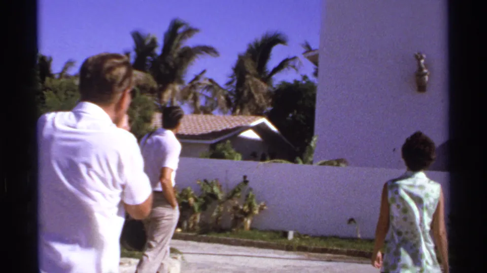 1974 Cabo San Lucas home-movie frame showing adults walking near a white stucco building, palms, and a tiled-roof structure.