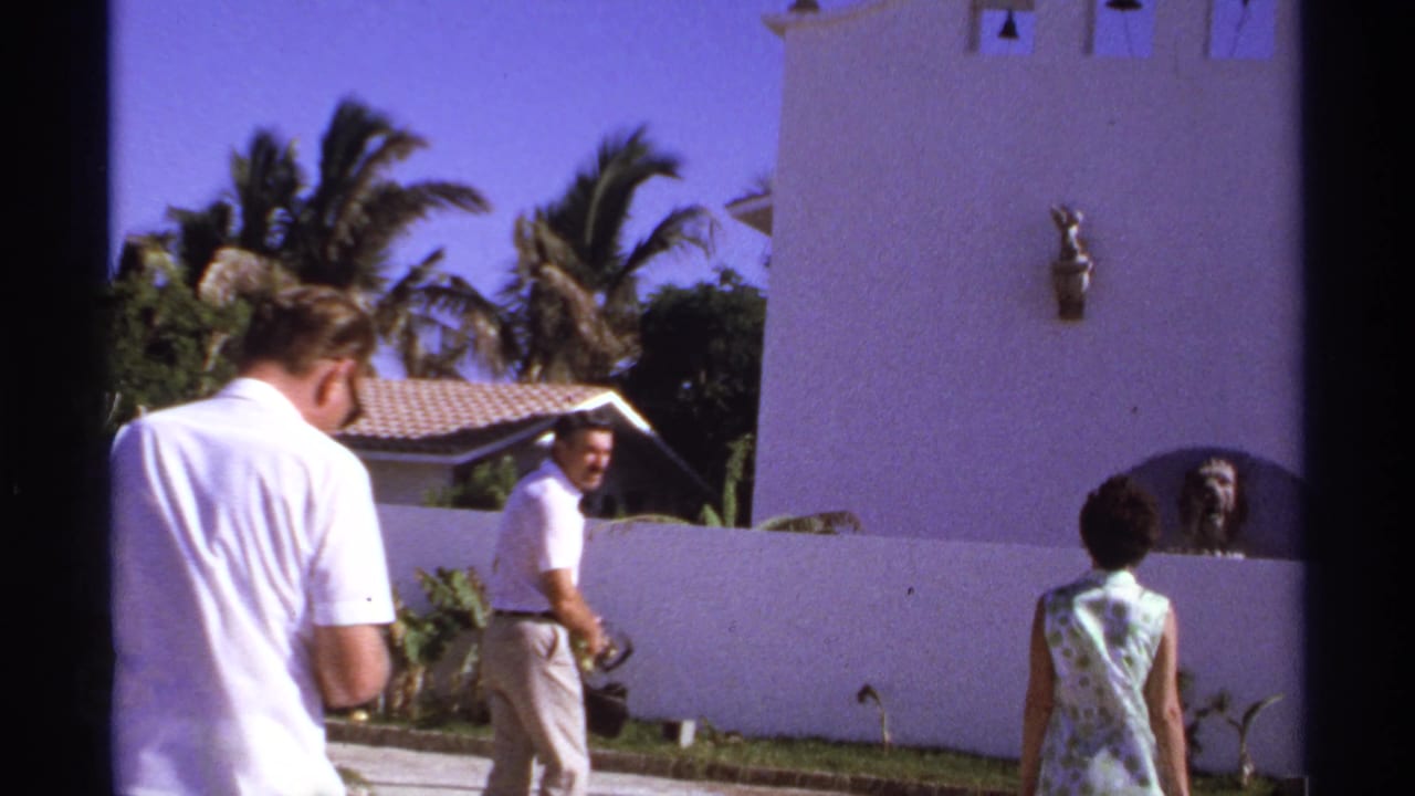 A man turns toward the camera as two other adults walk near a white bell-tower wall.