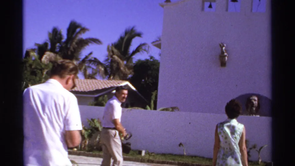 1974 Cabo San Lucas frame with a man glancing back, another man and woman walking, and bells on a white stucco facade.