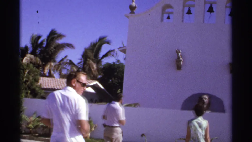 1974 Cabo San Lucas home-movie frame showing visitors near a white stucco facade with bells and a lion statue niche.