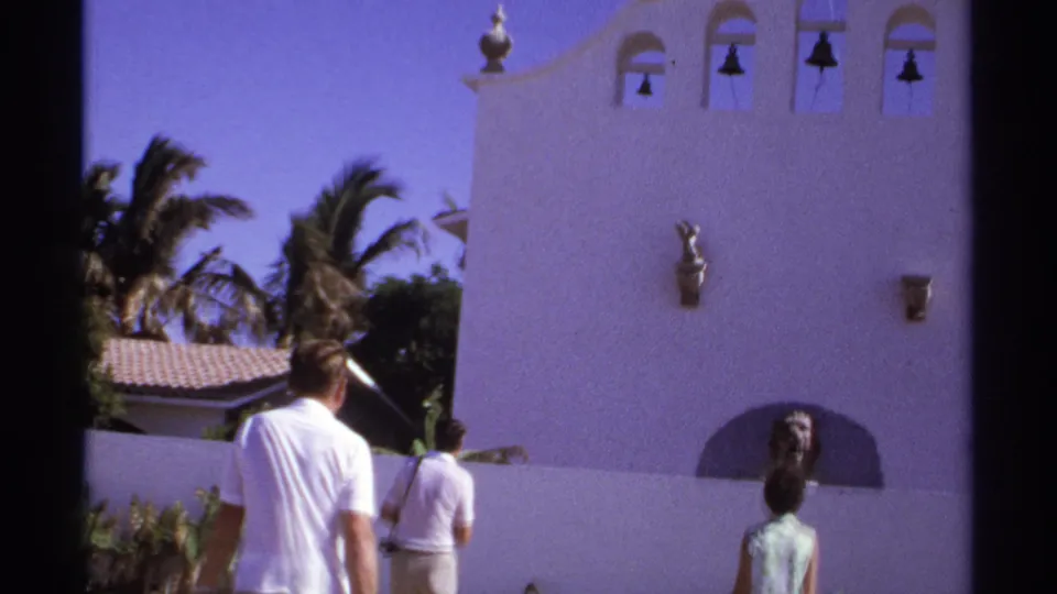 1974 Cabo San Lucas frame of three adult visitors walking below a white stucco bell facade with a lion statue niche.