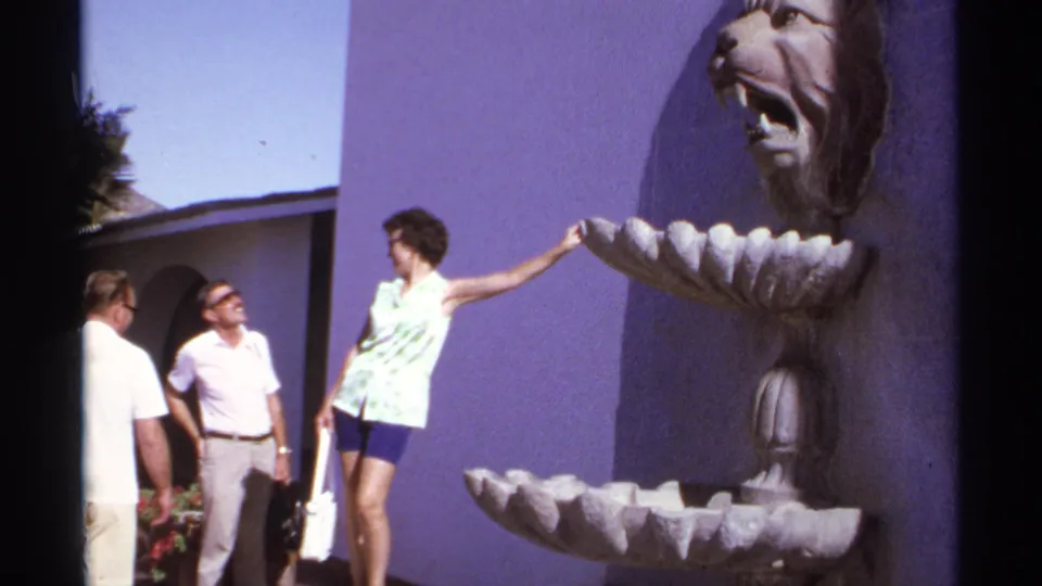 Cabo San Lucas 1974 frame showing two men and a woman near a lion-head fountain, with bright resort walls behind them.