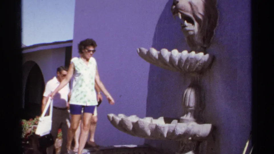 1974 Cabo San Lucas frame of a woman walking away from a decorative lion-head fountain as two men stand behind her.