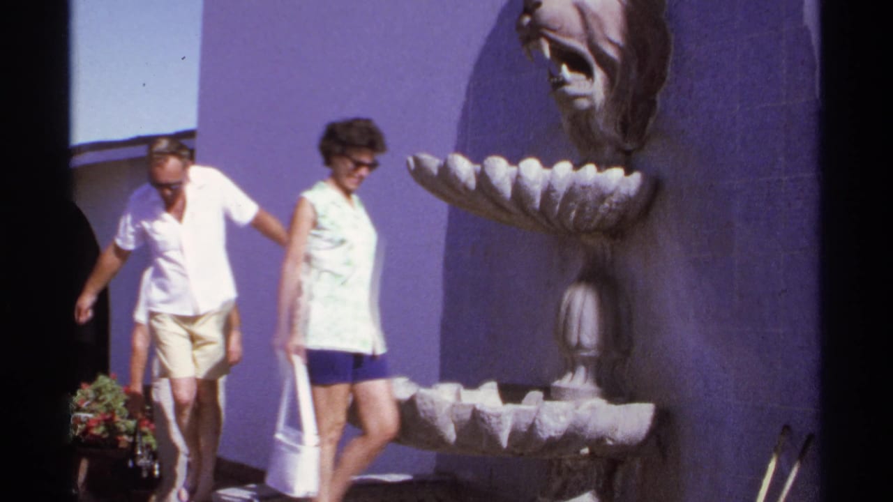 Two adults walk past a lion-head fountain beside a purple wall in bright sunlight.