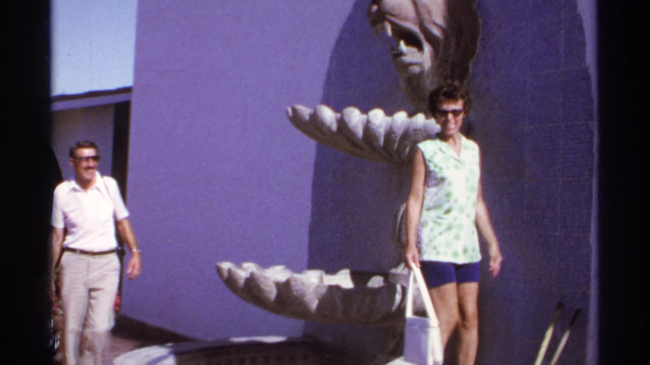 A woman walks near the fountain while a man follows beside the purple wall.