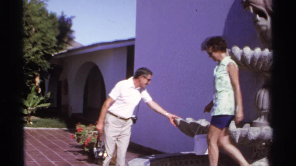 The man leans toward the stone fountain basin while the woman continues along the tiled courtyard path in 1974 Cabo San Lucas footage.