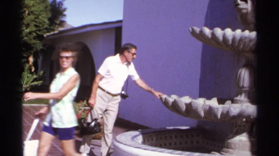 The woman moves past the camera with a tote bag as the man remains beside the stone fountain in the sunny Cabo San Lucas courtyard.