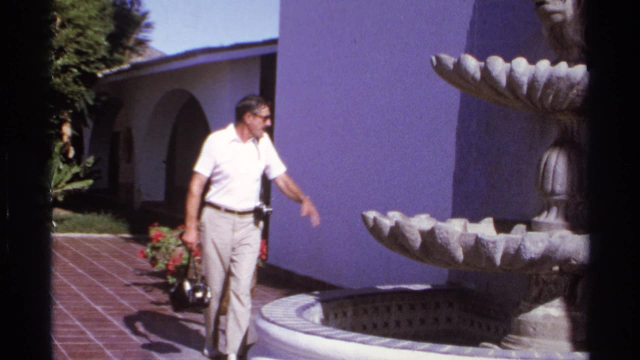 Man in a white polo walks beside a stone fountain in a sunny tiled courtyard.