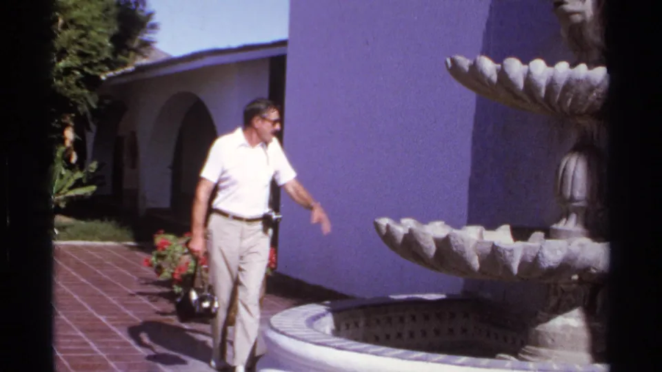 In 1974 Cabo San Lucas footage, an adult man with a camera walks toward a scalloped stone fountain in a sunlit courtyard.