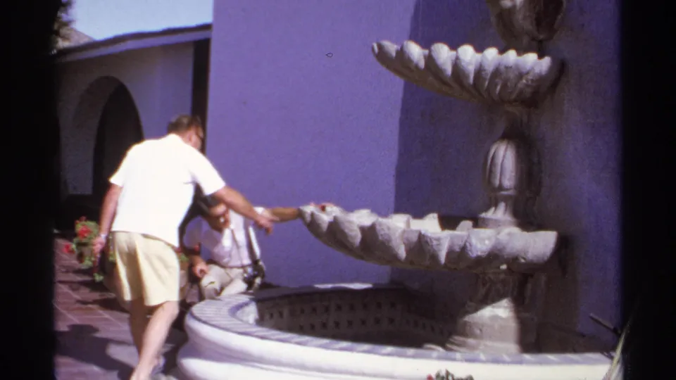 Two adult men appear beside the decorative fountain in 1974 Cabo San Lucas home-movie footage, one standing and one crouching.