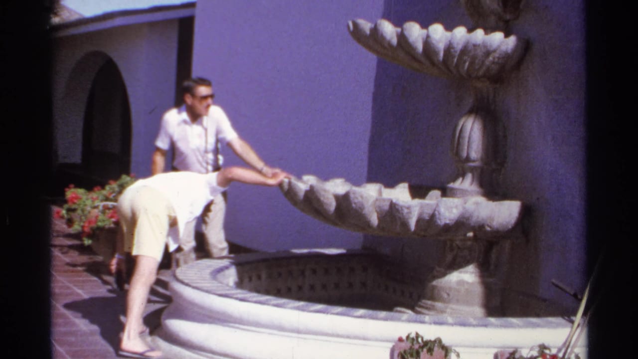 Man watches as another man leans over a tiered stone fountain in a sunny courtyard.