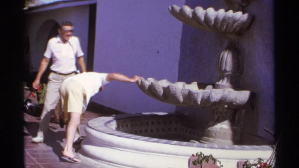 Two male tourists in 1974 Cabo San Lucas examine a large stone fountain set against a purple stucco courtyard wall.