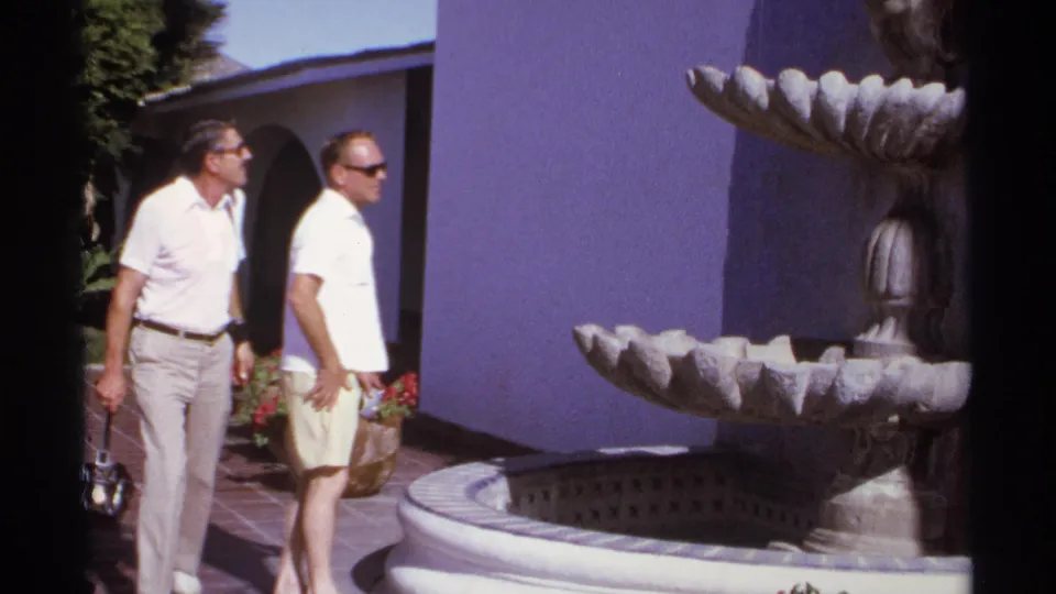 Two male tourists walk together beside the fountain in a Cabo San Lucas courtyard, with flowers and arched architecture visible.